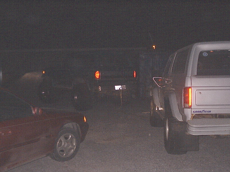 Several vehicles parked at night with taillights glowing, a light SUV at right