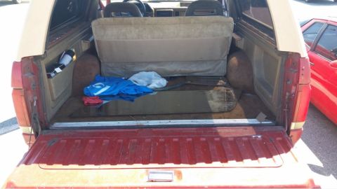 1989 Ford Bronco - The rear cargo area of a red and tan Ford Bronco shows a folded seat and various items, with a red car visible to the side.