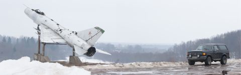 1987 Ford Bronco - A dark SUV is parked on a wet, snowy surface next to a white fighter jet on a pedestal.