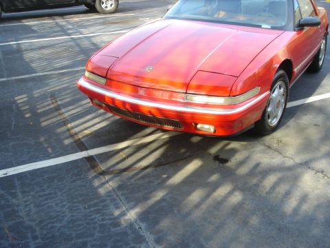 1980 Ford Mustang - This red coupe, likely a Buick Reatta, is parked in a lot with shadows cast across the asphalt.