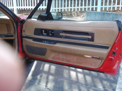 1980 Ford Mustang - The interior door panel of a red car features tan upholstery and wood-grain accents.