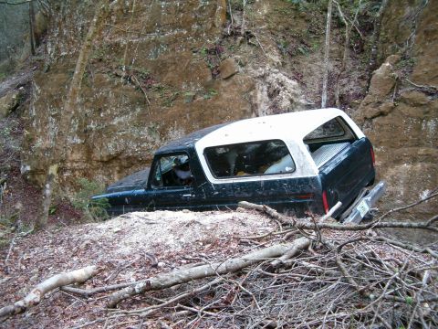 1991 Ford Bronco - Side view of a dark green Ford Bronco with a white removable hardtop leaned hard against a steep dirt-and-rock bank on a narrow off-road tr…