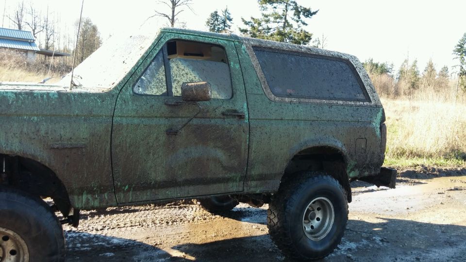 Side view of a green 1990 Ford Bronco covered in mud, parked on a dirt road