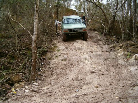 1991 Ford Bronco - This green Ford Bronco is navigating a steep, muddy incline in a wooded area. The windshield displays the website 'FULLSIZEBRONCO.COM'.