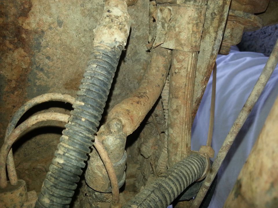 Corroded metal pipes and black corrugated hoses under a vehicle, with a blue cloth in the background