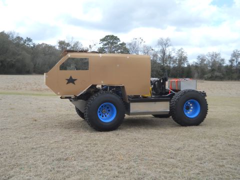 1992 Ford Bronco - A custom-built vehicle with large tires and a boxy tan body sits in a grassy field under a cloudy sky.