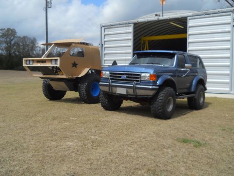 1992 Ford Bronco - Two modified SUVs, a 1992 Ford Bronco and a custom build, sit in a grassy lot near a large metal structure.