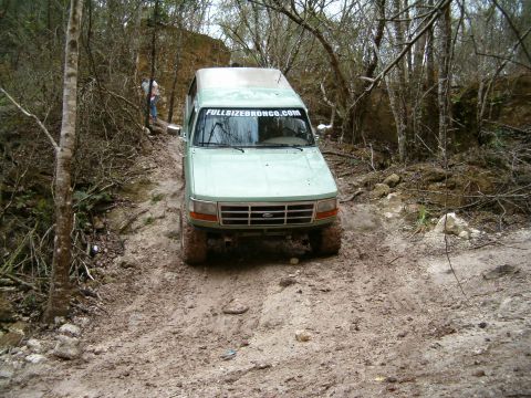1991 Ford Bronco - A light green Ford Bronco navigates a challenging muddy path in a wooded area.