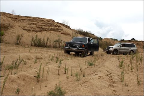 1987 Ford Bronco - Two SUVs navigate a sandy landscape with sparse vegetation and rolling dunes.