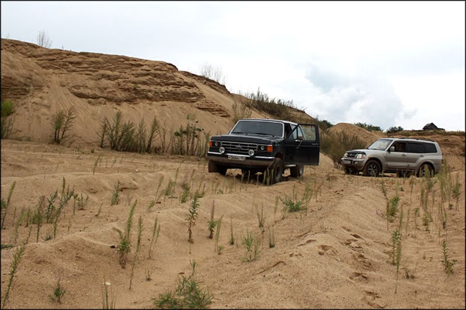 Black SUV with door open and silver SUV on sandy terrain under a cloudy sky