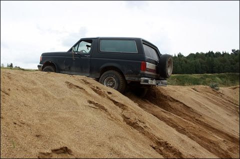1987 Ford Bronco - A black 1987 Ford Bronco navigates a sandy incline under a cloudy sky.