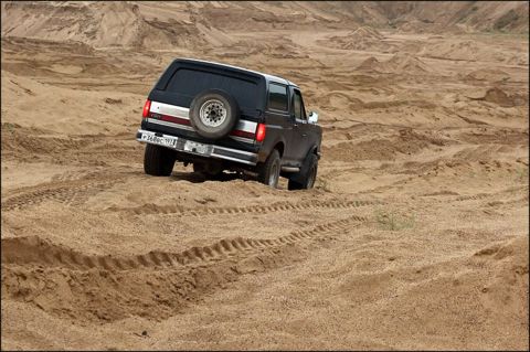 1987 Ford Bronco - 1987 Ford Bronco navigates sandy terrain, showing its rear profile and spare tire.