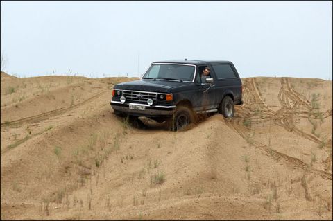 1987 Ford Bronco - A black Ford Bronco navigates sandy terrain, kicking up dust as it ascends a dune.
