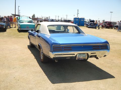 1967 Pontiac GTO - Rear view of a blue 1967 Pontiac GTO with a white vinyl top, parked outdoors with other vehicles and people around.