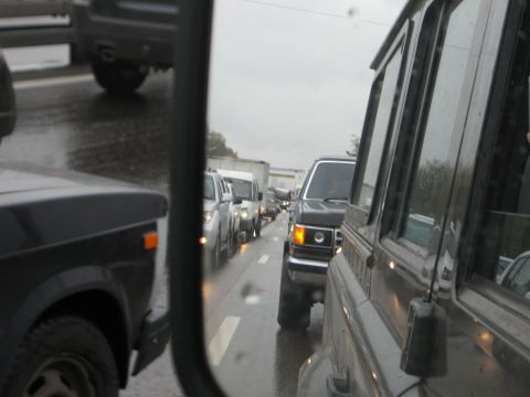 1987 Ford Bronco - A line of vehicles is stuck in traffic on a rainy day, as seen from the driver's side mirror.