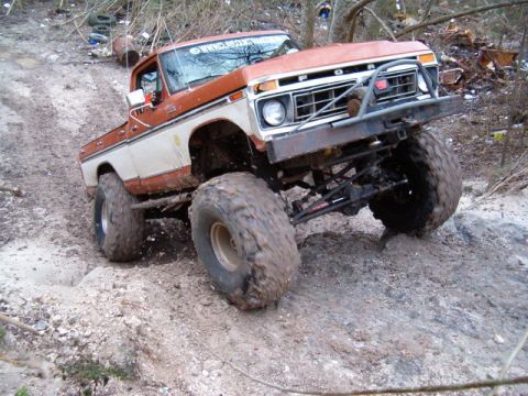 1991 Ford Bronco - Front three-quarter view of a lifted orange-and-white 1970s Ford F-series 4x4 flexing on a rocky clay trail, showing exposed leaf-sprung so…