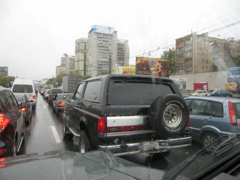 Black Ford Bronco with spare tire on back, stuck in traffic on a wet day. Other cars and buildings visible