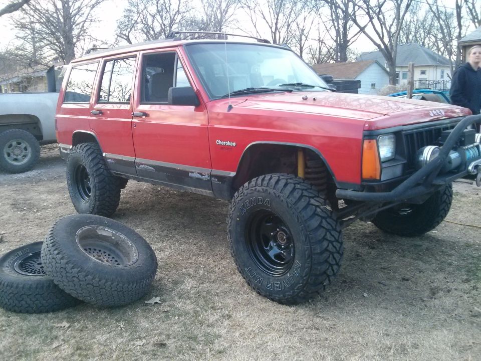 Red Jeep Cherokee with aftermarket bumper and winch, two spare tires on the ground
