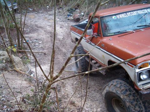 1991 Ford Bronco - A lifted Ford pickup truck navigates a muddy, wooded area with debris scattered around.