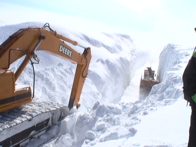 Yellow Deere excavator in foreground, orange bulldozer in background clearing a path through deep snow