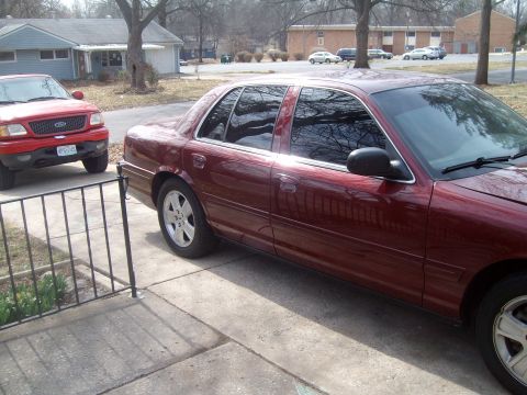2004 Ford Crown Victoria - A red sedan is parked in a driveway next to a red pickup truck on a sunny day.