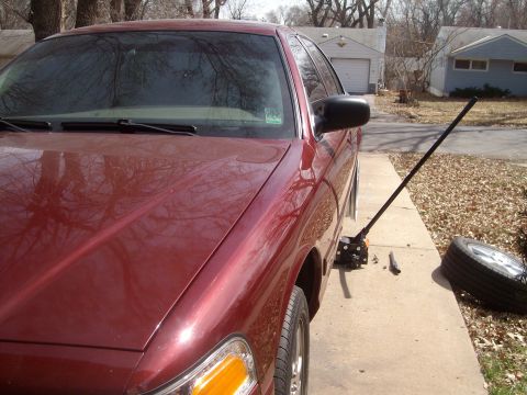 2004 Ford Crown Victoria - A red 2004 Ford Crown Victoria is parked on a driveway with a jack positioned to lift the front passenger side.