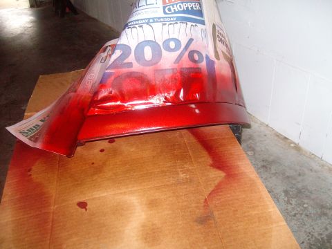 2004 Ford Crown Victoria - A red car bumper is being painted, with newspaper and paint drips visible on the cardboard surface.