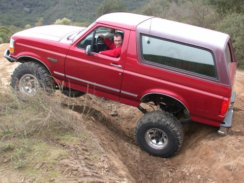 1996 Ford Bronco - A man drives a red Ford Bronco through a challenging off-road terrain, with one tire lifted high.