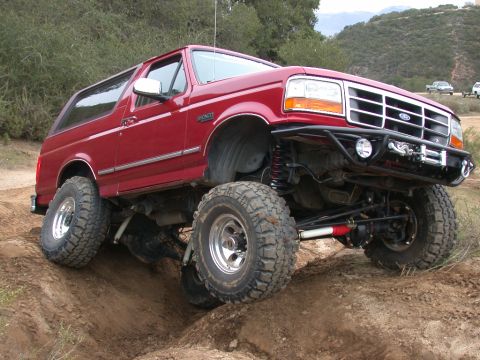1996 Ford Bronco - A red 1996 Ford Bronco is shown off-roading on a dirt path with hills in the background.