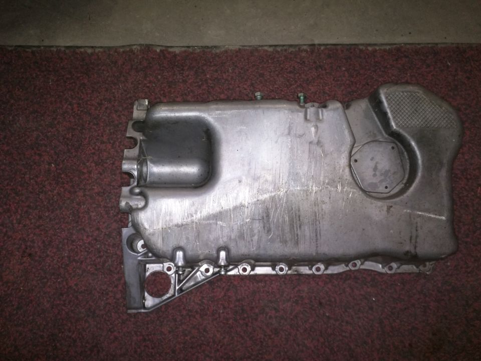 Metal oil pan with mounting holes and a hexagonal cap, resting on a red carpet