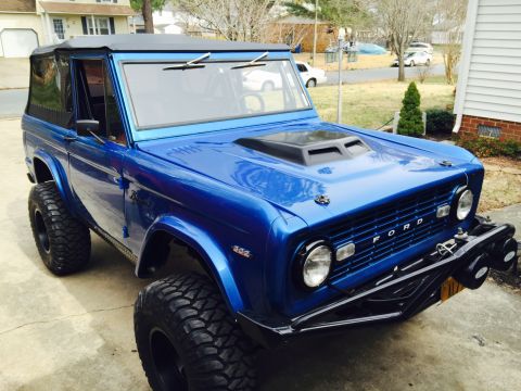 1974 Ford Bronco - This blue 1974 Ford Bronco features a black soft top, a prominent aftermarket hood scoop, and aggressive off-road tires.