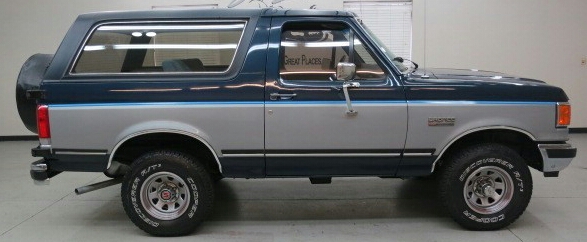 Two-tone blue and silver Ford Bronco with a spare tire on the rear, seen from the side