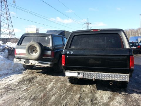 1987 Ford Bronco - Two Ford Broncos are parked side-by-side on a snowy, unpaved road under a bright blue sky.