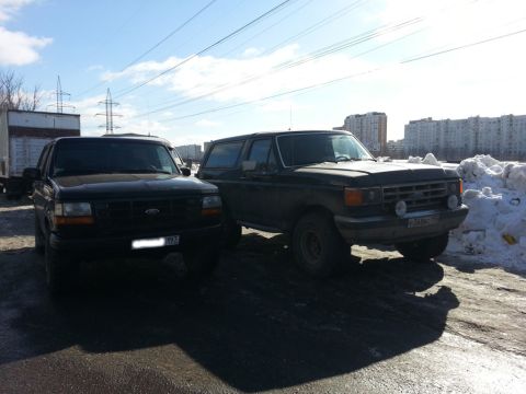 1987 Ford Bronco - Two Ford Broncos, a dark green 1987 model and a black later model, are parked side-by-side on a sunny day.