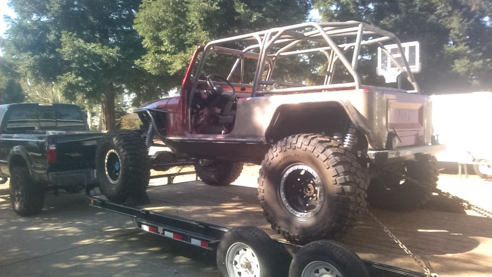 Modified Jeep CJ-series on a trailer, red body, roll cage, large tires, being towed by a black pickup truck