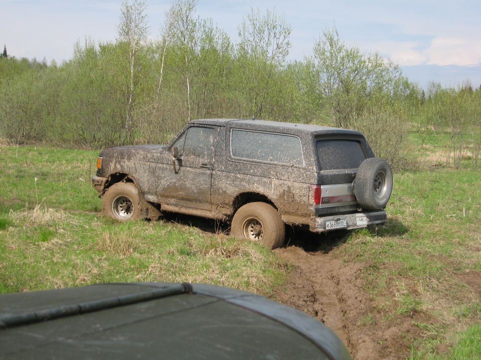 Muddy 1987 Ford Bronco stuck in a muddy field, with another vehicle's hood visible in the foreground
