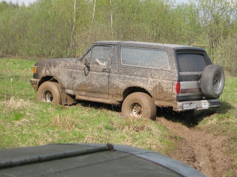 1987 Ford Bronco - A muddy 1987 Ford Bronco is stuck in a muddy field, with another vehicle partially visible in the foreground.
