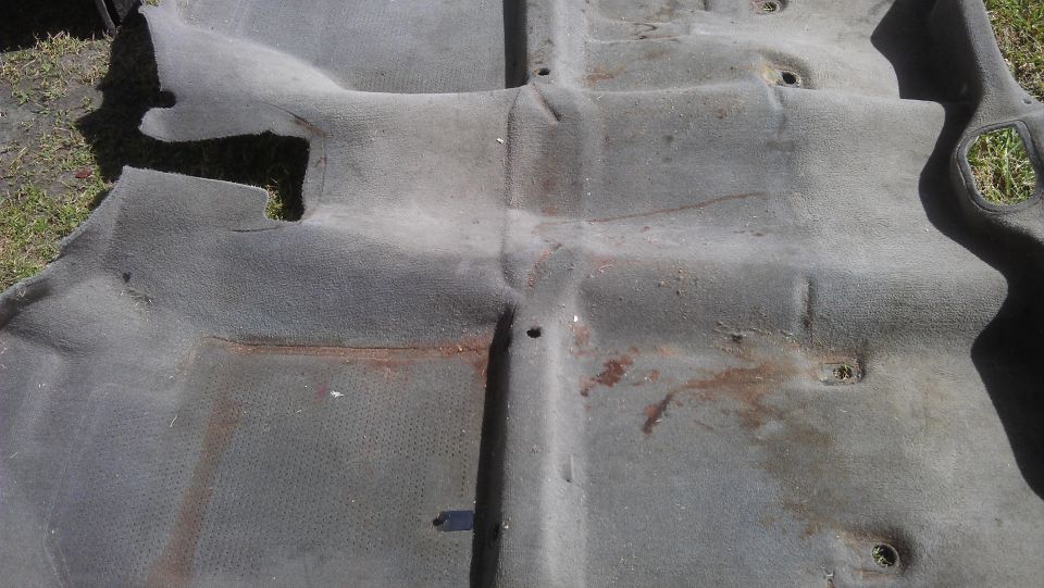 Worn gray car carpet with rust stains and visible floor pan indentations