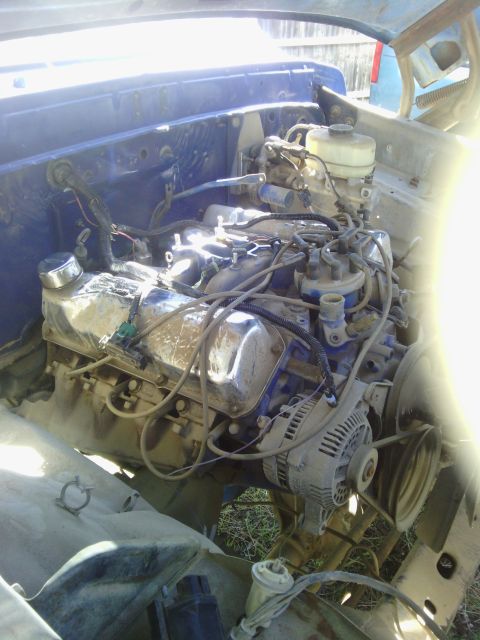 1978 Ford Bronco - The engine bay of a vehicle, showing a V8 engine, alternator, and various hoses and wires. The firewall is painted blue.