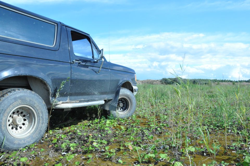 1987 Ford Bronco parked in a muddy field with tall grass and a blue sky