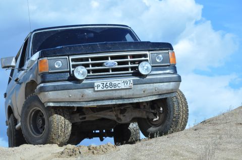 1987 Ford Bronco - This Ford Bronco is positioned on a sandy hill with a bright blue sky and clouds above.