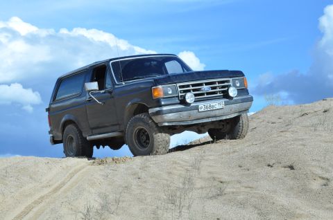 1987 Ford Bronco - A black 1987 Ford Bronco ascends a sandy incline, its tires kicking up dust against a backdrop of blue sky and scattered clouds.