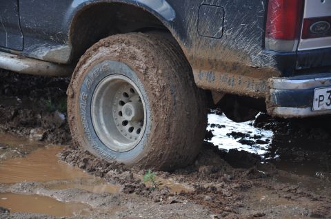 1987 Ford Bronco - A 1987 Ford Bronco's rear tire is deeply stuck in mud, with a Ford badge visible on the tailgate.