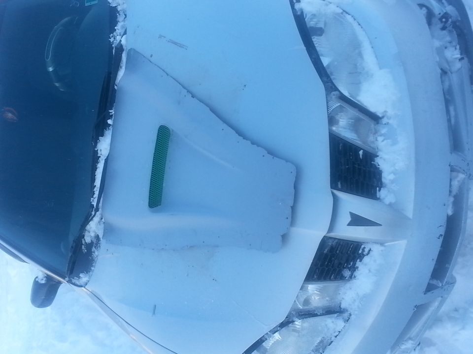 White car hood covered in snow, with a green vent and a black grille visible