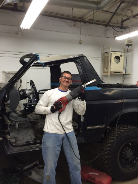 1992 Ford Bronco - A man works on modifying the roof of a dark SUV, likely a 1992 Ford Bronco, with a reciprocating saw.
