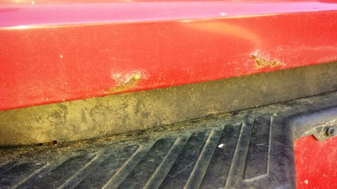 2001 Ford F-150 - Close-up of a red truck bed corner showing rust damage and black plastic trim, consistent with a 2001 Ford F-150.
