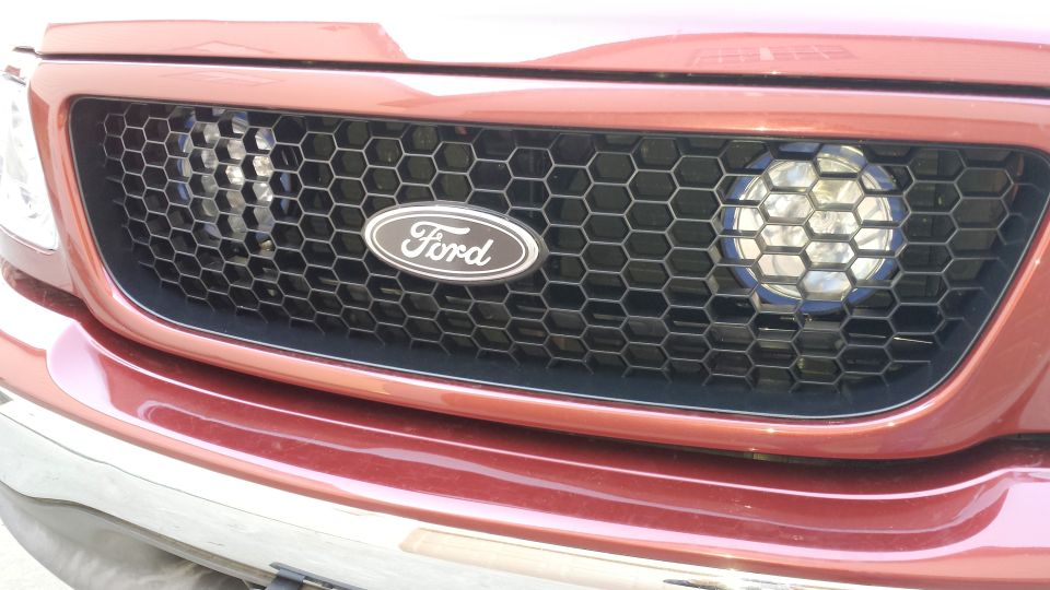 Close-up of a 2001 Ford F-150 grille with a honeycomb pattern and a Ford oval badge. A round headlight is visible to the right