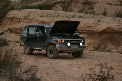 1987 Ford Bronco - A black Ford Bronco sits on a dirt road with its hood open, surrounded by sandy terrain.