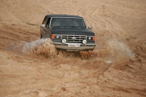 1987 Ford Bronco - A 1987 Ford Bronco navigates a muddy terrain, kicking up water and dirt.