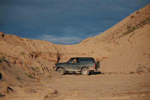 1987 Ford Bronco - A dark 1987 Ford Bronco is parked on a sandy hill under a cloudy blue sky.